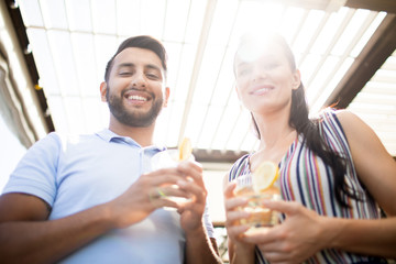 Young smiling man and his girlfriend with drinks looking at camera on sunny day while enjoying their date