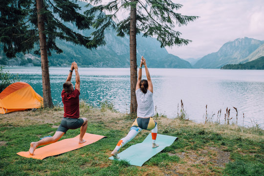 Back view of sporty couple near orange tent with their hands apart against the background of lake and mountains. The man and woman workout yoga with mats at camping in summer norwegian cloudy morning. - Powered by Adobe