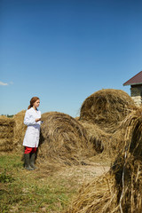 Naklejka na ściany i meble Young woman in whitecoat standing by stack of hay while browsing in the net