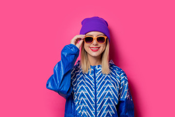 Young blonde girl in 90s sports jacket and hat with sunglasses on pink background.