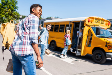 rear view of smiling teen african american schoolboy walking to school bus with his classmates