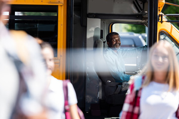 happy mature school bus driver looking at teenagers walking on foreground