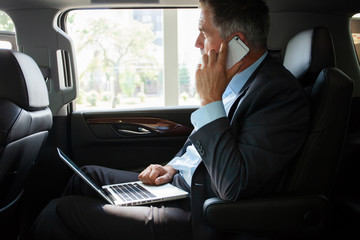Handsome businessman talking with phone sitting with laptop on the backseat of the car.