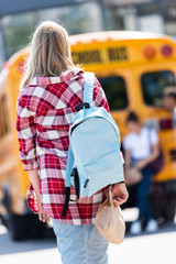 rear view of teen schoolgirl with backpack walking to classmates leaning on school bus