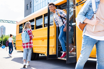 smiling african american teen schoolboy walking out of school bus wit classmates around