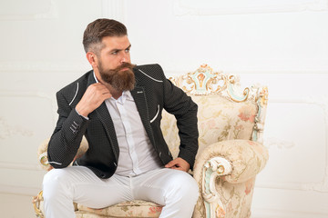 Imposing well dressed man sitting on couch. Men's beauty, fashion. Cool man in formal wear, tux pocket square, sit in chair over white background, looking away, shares, stock, money, rich, brutal.