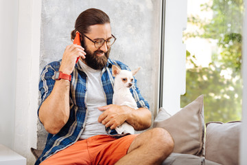Looking at dog. Bearded caring owner of dog wearing squared shirt looking at him speaking on phone