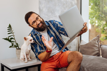 Video chat. Dark-haired beaming man feeling emotional while having video chat sitting near dog