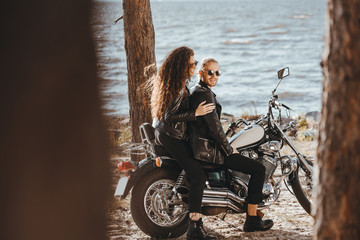 young couple of bikers in black leather jackets sitting on chopper motorcycle on seashore