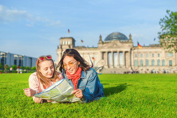 Obraz na płótnie Canvas Multiracial group of friends visiting the city of Berlin. Two women reading map with Reichstag building on the background. Friendship and travel concept with real candid emotions