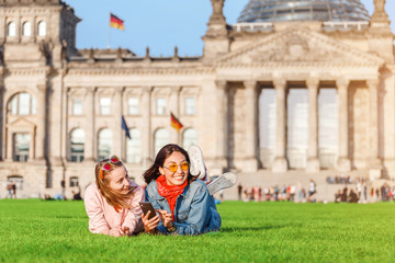 Obraz na płótnie Canvas Two Young happy girls wearing sun glasses lying on a grass and have fun in front of the Bundestag building in Berlin. Studying abroad and travel in Germany concept