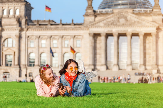 Two Young happy girls wearing sun glasses lying on a grass and have fun in front of the Bundestag building in Berlin. Studying abroad and travel in Germany concept