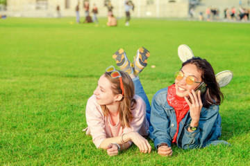 Two student girls lying on a green grass and talking by mobile phone