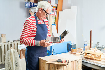 Side view of elderly man in apron looking at laptop screen while working at carpenter workshop