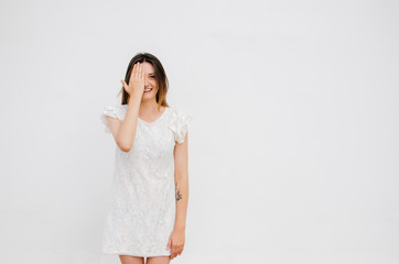 Smiling woman, with eye, clossed by hand in white dress on white wall