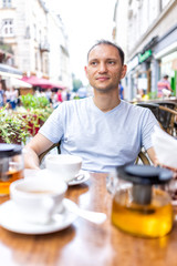 Fototapeta na wymiar Young happy man sitting in European outdoor cafe restaurant looking people watching drinking tea with glass teapot, cups on romantic date in summer in Lviv, Ukraine