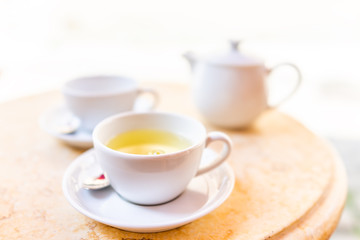 Closeup of two white cups on plates of green or oolong tea in breakfast brunch outdoor cafe restaurant outside wooden table with sunny colorful sunlight, teapot, pot