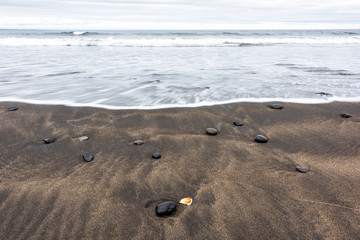 Closeup of large black volcanic rocks on sand beach in north west Iceland, with long exposure smooth water waves on shore, shiny wet stones, orange sea shell seashell, Snaefellsnes peninsula