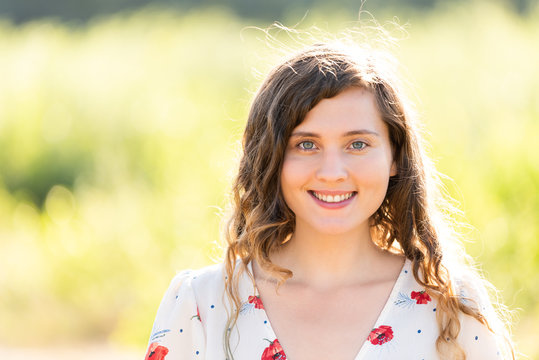 Closeup portrait of young woman's face smiling happy in sunny summer green park at sunset showing teeth, wavy hair in wind, bokeh background