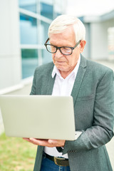 Senior man in glasses using modern laptop while standing on blurred background of building