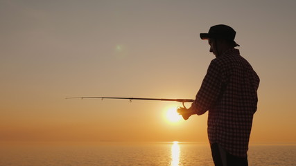 Silhouette of a young fisherman fishing on the beach at sunset. Side view