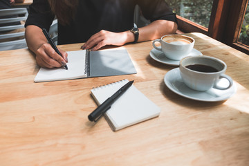 Closeup image of a woman's hand writing down on a white blank notebook with coffee cup on wooden table