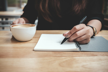 Fototapeta na wymiar Closeup image of a woman's hand writing down on a white blank notebook while drinking coffee on wooden table