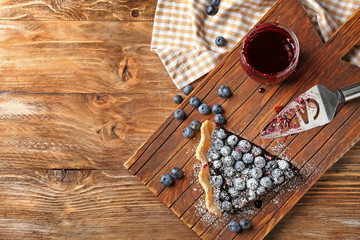 Wooden board with piece of delicious blueberry pie and jar of jam on table