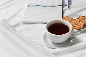 Cup of tea, cookies and notebook on bed linen