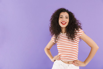 Photo of pretty hipster woman with curly hair in summer wear smiling and looking at you, isolated over violet background in studio