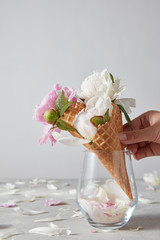 Greeting card womans hand takes a wafer cone with tender white peony from glass vase. Petals of flowers on a gray stone table.