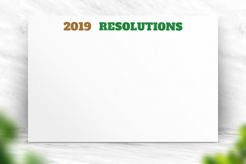 2019 new year resolutions text on white paper poster with natural blur leaf foreground on white marble room wall,Business presentation mock up for adding your list.