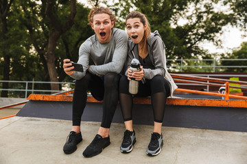 Fototapeta premium Image of excited or shocked man and woman 20s in sportswear, sitting on parapet in green park after training or fitness on sunny summer day