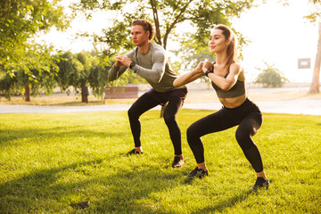 Image of young caucasian sporty man and woman 20s in tracksuits, doing workout and squatting...