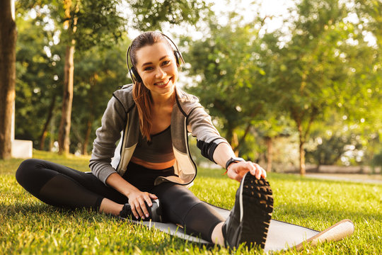 Photo of athletic sportswoman 20s in sportswear listening to music and stretching foot on grass, during workout in park