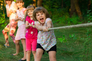 Group of happy children playing tug of war outside on grass. Kids pulling rope at park. Summer camp...