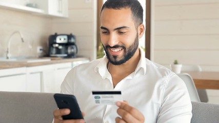 Happy handsome man making purchase online, typing credit card details on black smartphone, indoor shot in modern studio apartment at the weekend