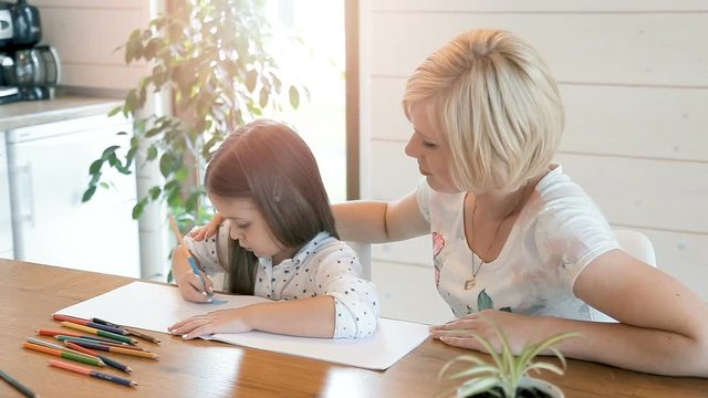 Caring mother helping her school child to draw, beautiful blond woman sitting next to cute long-haired kid in large light kitchen