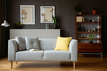 Yellow pillow on grey couch in vintage living room interior with lamp and posters. Real photo