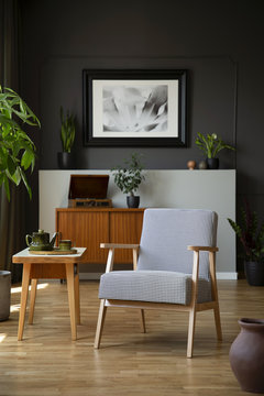 Patterned armchair next to wooden table in dark grey living room interior with poster. Real photo