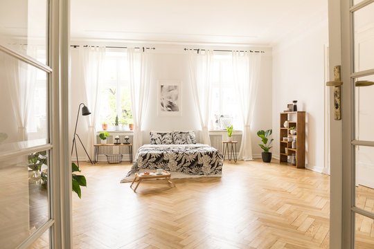 View through an open double door into a bedroom interior with wooden parquet, large windows and white walls. Bed with linen, breakfast tray, lamp and bookcase in the room. Real photo.