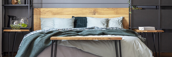 Panorama of wooden bench in front of bed with green blanket in grey bedroom interior. Real photo