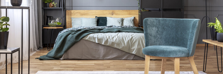 Blue armchair in grey bedroom interior with green blanket on wooden bed. Real photo