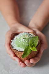 Woman hands hold frozen green sorbet in the coconut shell with fresh mint leaf on gray stone...