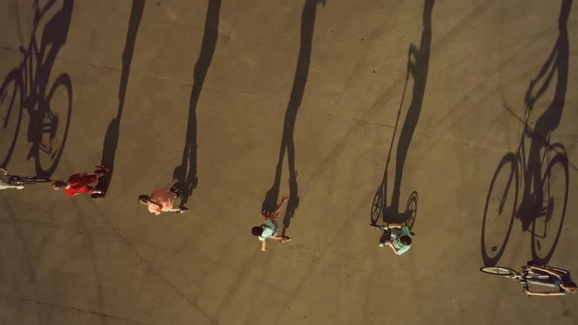 Teenagers riding in row on bikes, roller skates and scooters cast long shadows. Top view