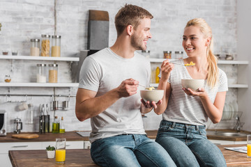 cheerful couple holding plates and spoons with corn flakes and looking at each other in kitchen