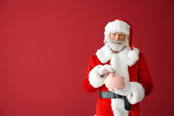Santa Claus putting coin into piggy bank on color background