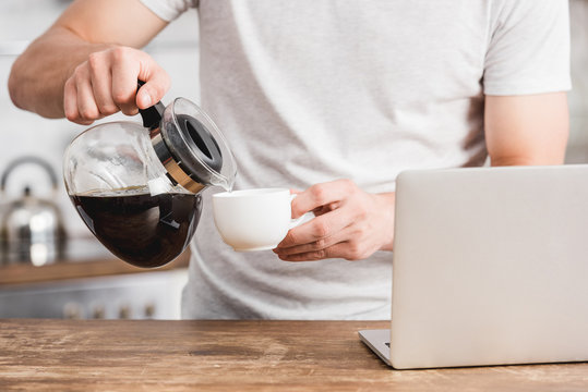 cropped image of man pouring coffee into cup near laptop in kitchen