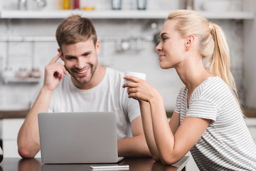 smiling couple sitting in kitchen with laptop and cup of coffee