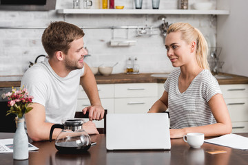 smiling young couple talking and looking at each other while using laptop together at home